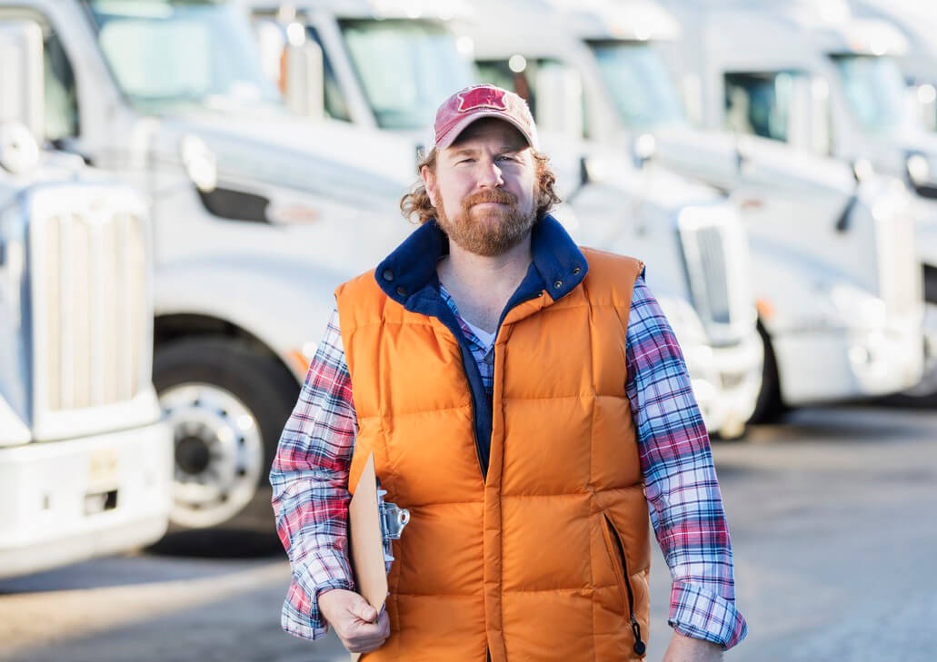 A mature man in h is 40s standing in front of a fleet of semi-trucks or tractor-trailers, holding a clipboard, with a serious, confident expression. He is an experienced truck driver or the owner of manager of a trucking company.